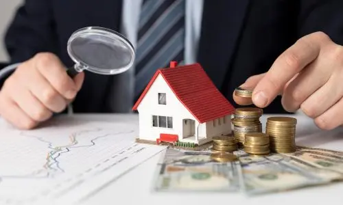 A person analyzing finances and property value with a magnifying glass, coins, and a house model