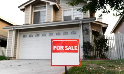 suburban house with a "For Sale" sign in front. The house has a two-car garage, multiple windows, and appears to be two stories tall